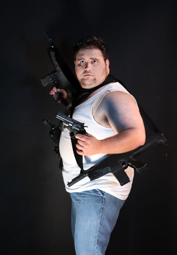 Actor in dirty white tank and jeans holding lots of gunes